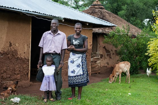 A man and woman and their child standing in front of their home and a cow